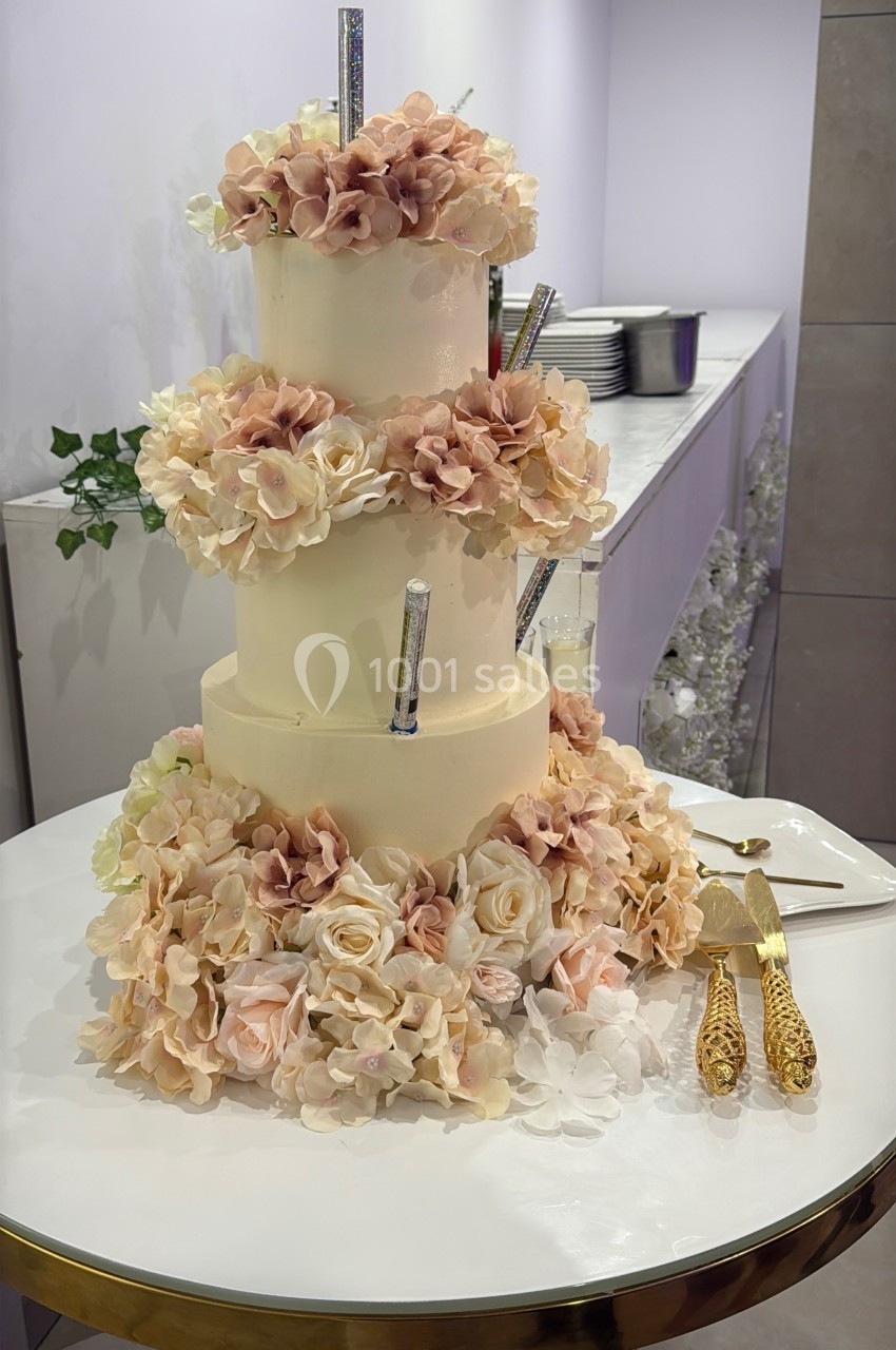 Gâteau de mariage à trois étages décoré de fleurs pastel et de bougies scintillantes, posé sur une table blanche.