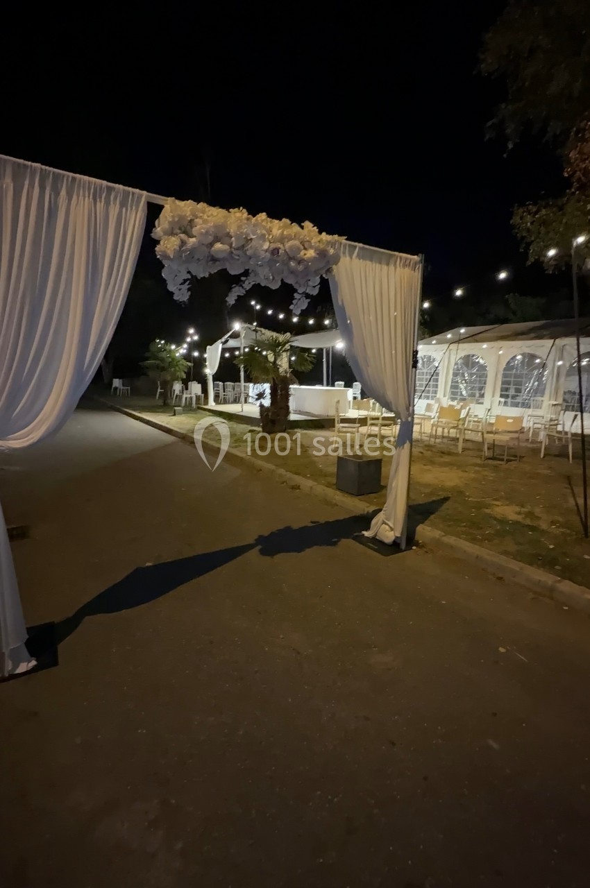 Arche décorée de rideaux blancs et de fleurs, éclairée par des guirlandes lumineuses dans un espace extérieur nocturne.