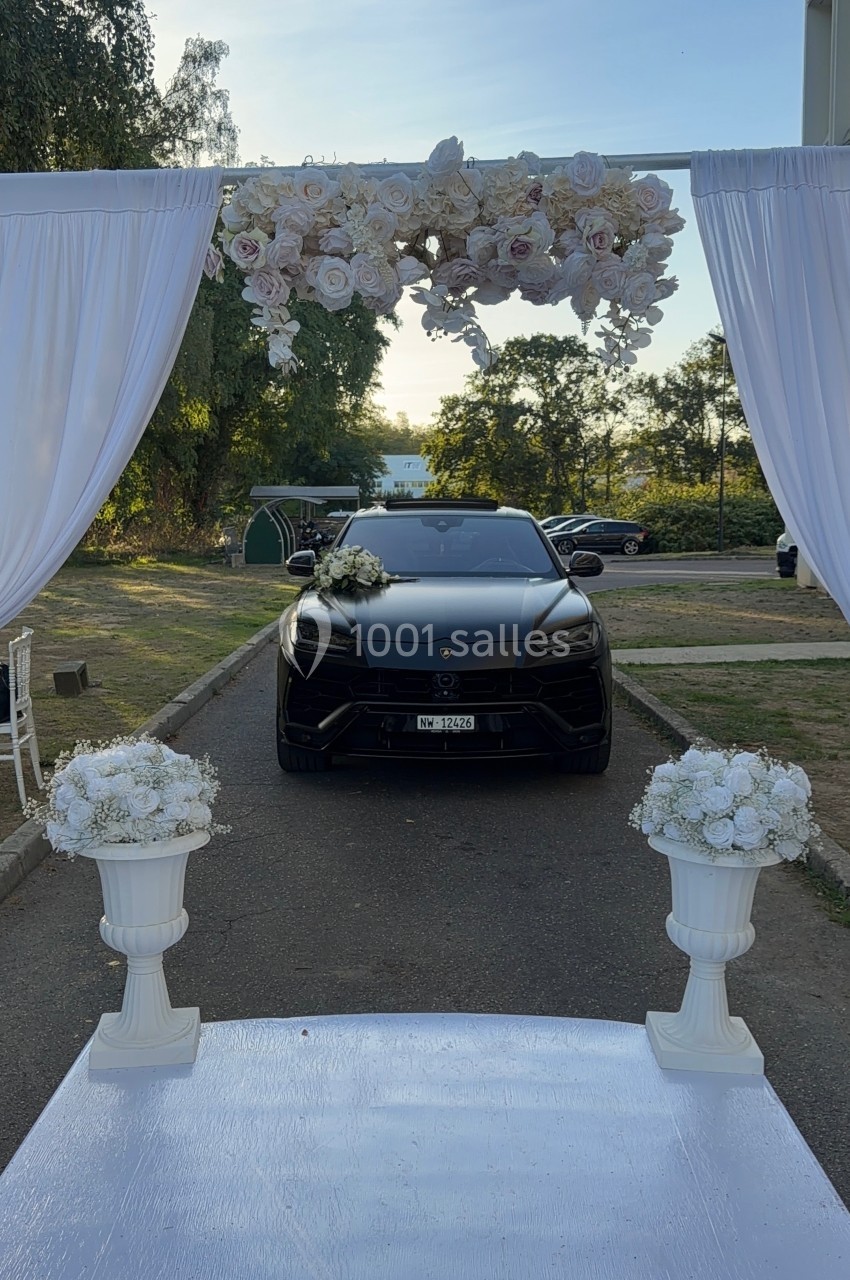 Voiture noire décorée de fleurs blanches, stationnée sous une arche ornée de rideaux et de fleurs blanches.