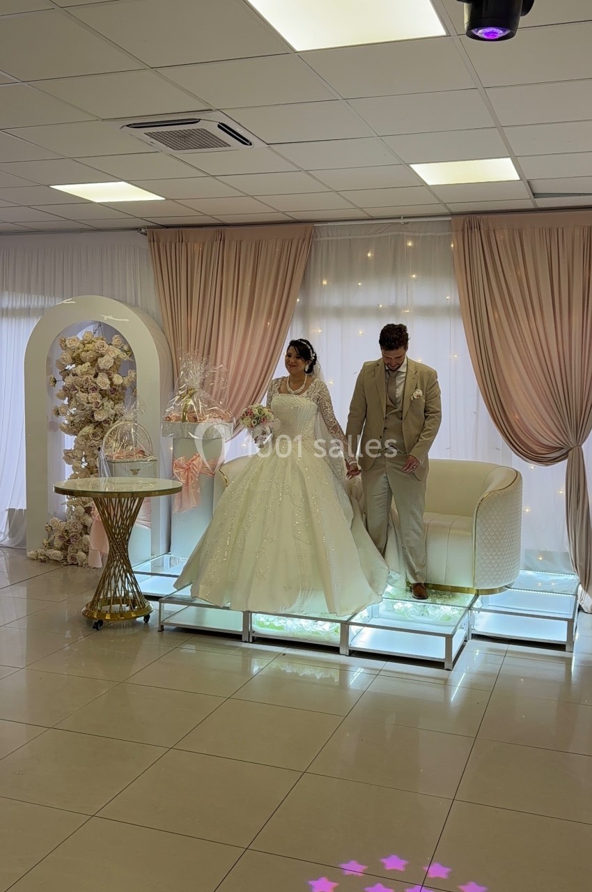 Un couple en tenue de mariage pose sur une estrade décorée avec des rideaux roses et des fleurs.