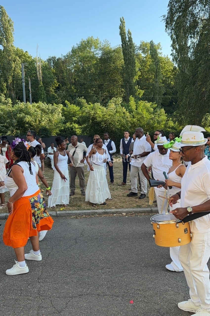 Un groupe de personnes danse et joue de la musique en plein air, entouré de végétation et de spectateurs.