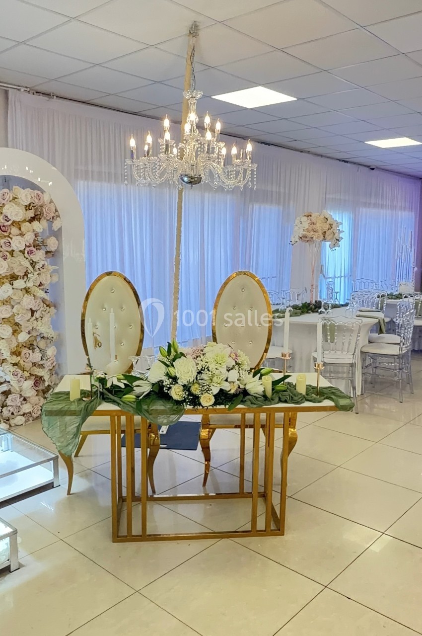 Table décorée pour un mariage avec fleurs blanches, chandeliers et fauteuils, dans une salle lumineuse ornée de rideaux…