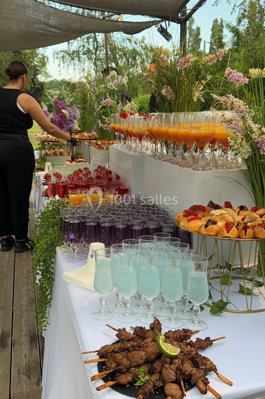 Buffet en plein air avec cocktails colorés, brochettes, viennoiseries et décorations florales sur une table blanche.