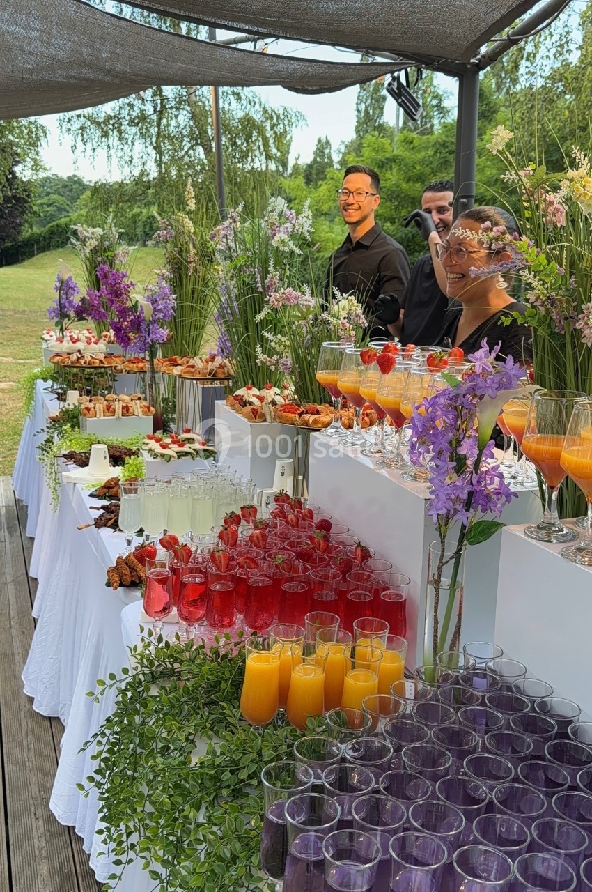 Buffet en plein air avec boissons colorées, fleurs décoratives et serveurs en arrière-plan sous une pergola.