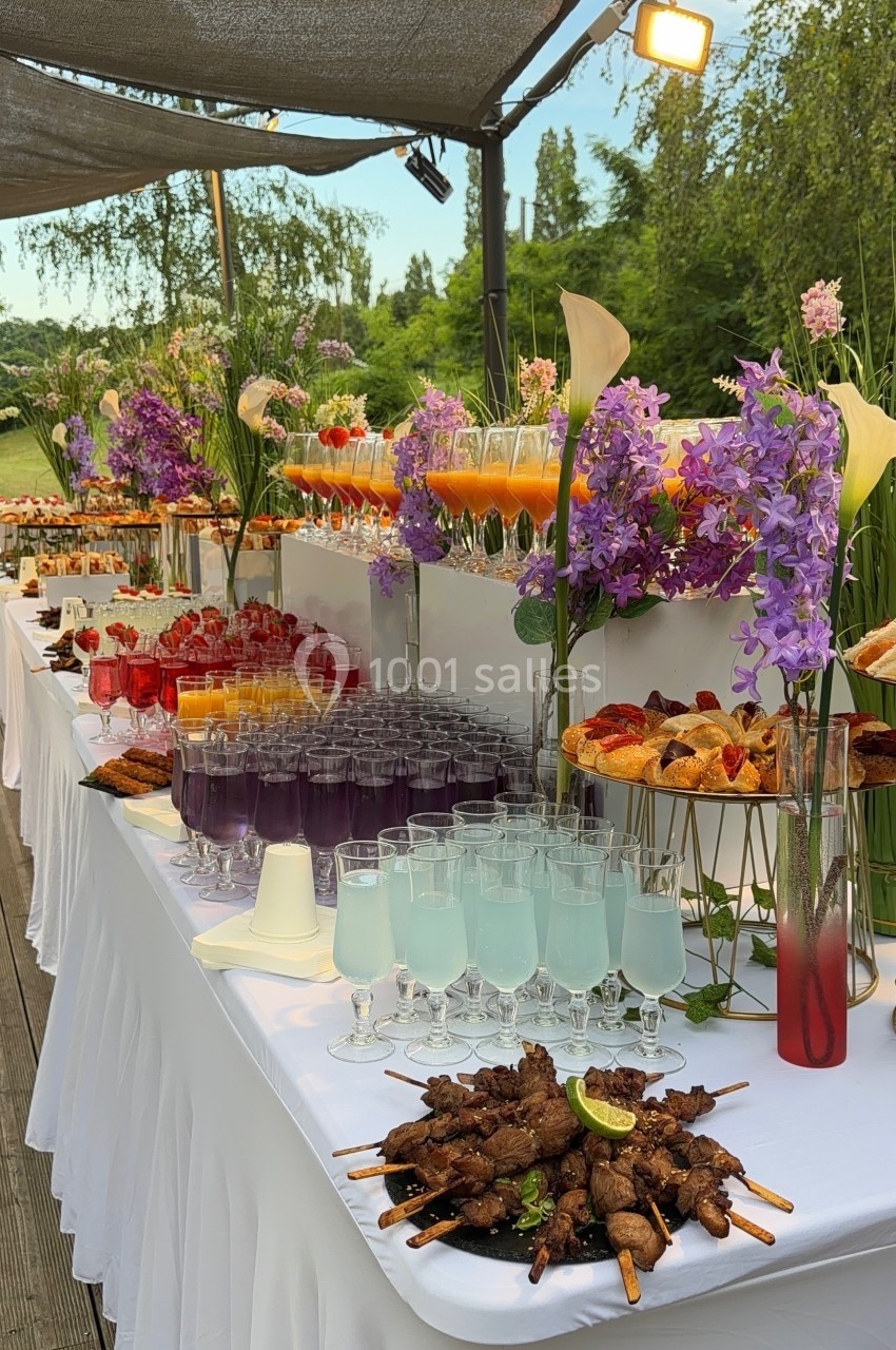 Buffet extérieur avec cocktails colorés, brochettes, viennoiseries et décorations florales sous une pergola.
