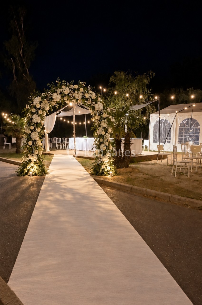 Allée blanche menant à une arche fleurie éclairée, avec des guirlandes lumineuses et des tentes en arrière-plan.