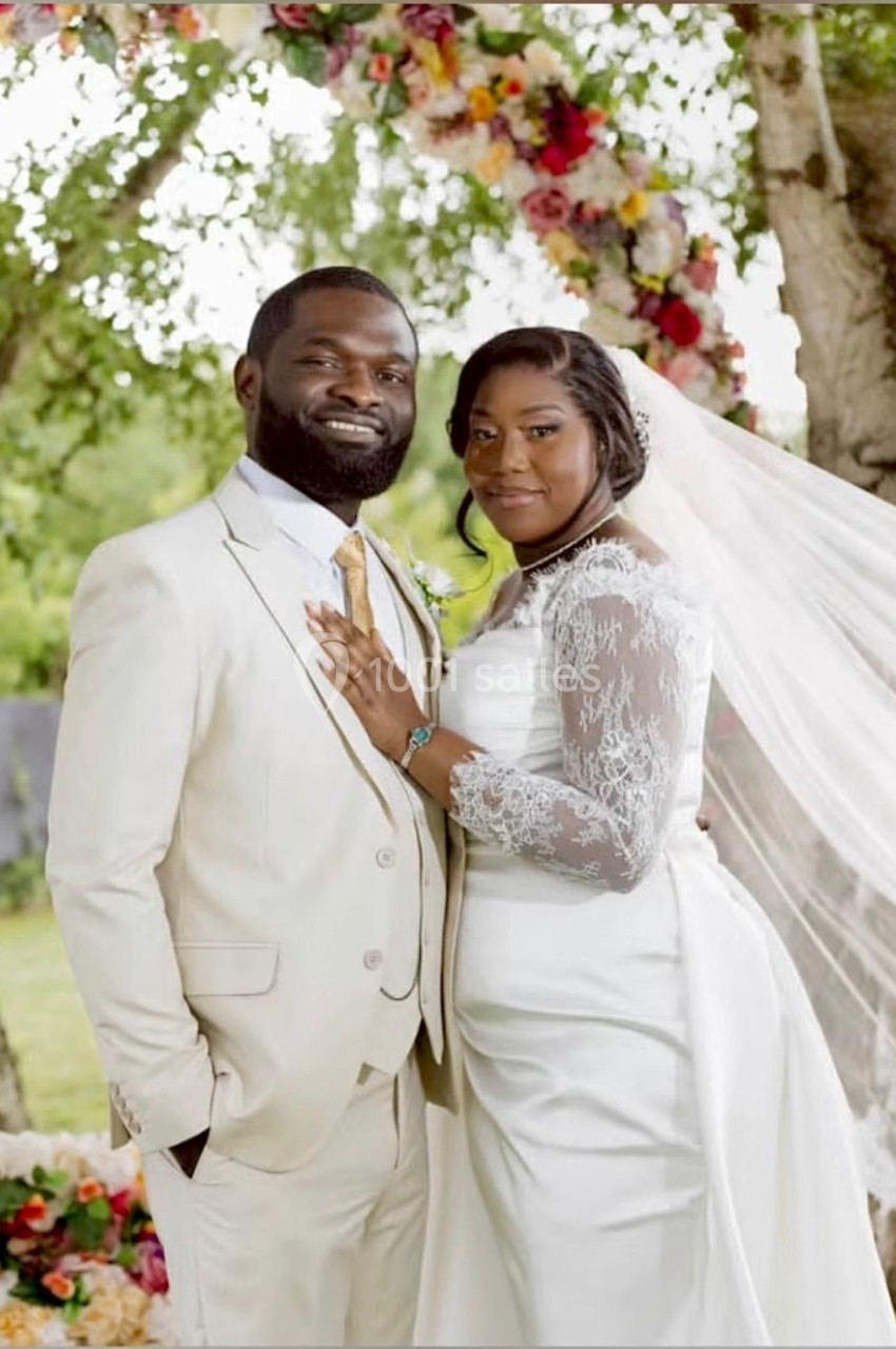 Un couple en tenue de mariage pose devant une arche fleurie dans un cadre extérieur verdoyant.