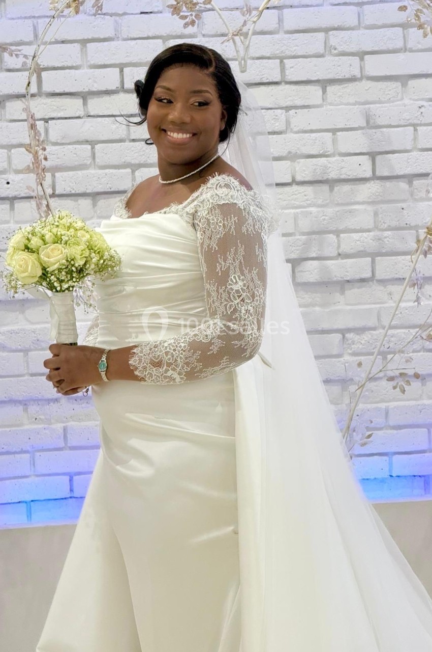 Femme en robe de mariée blanche tenant un bouquet de fleurs, posant devant un mur en briques blanches décoré.