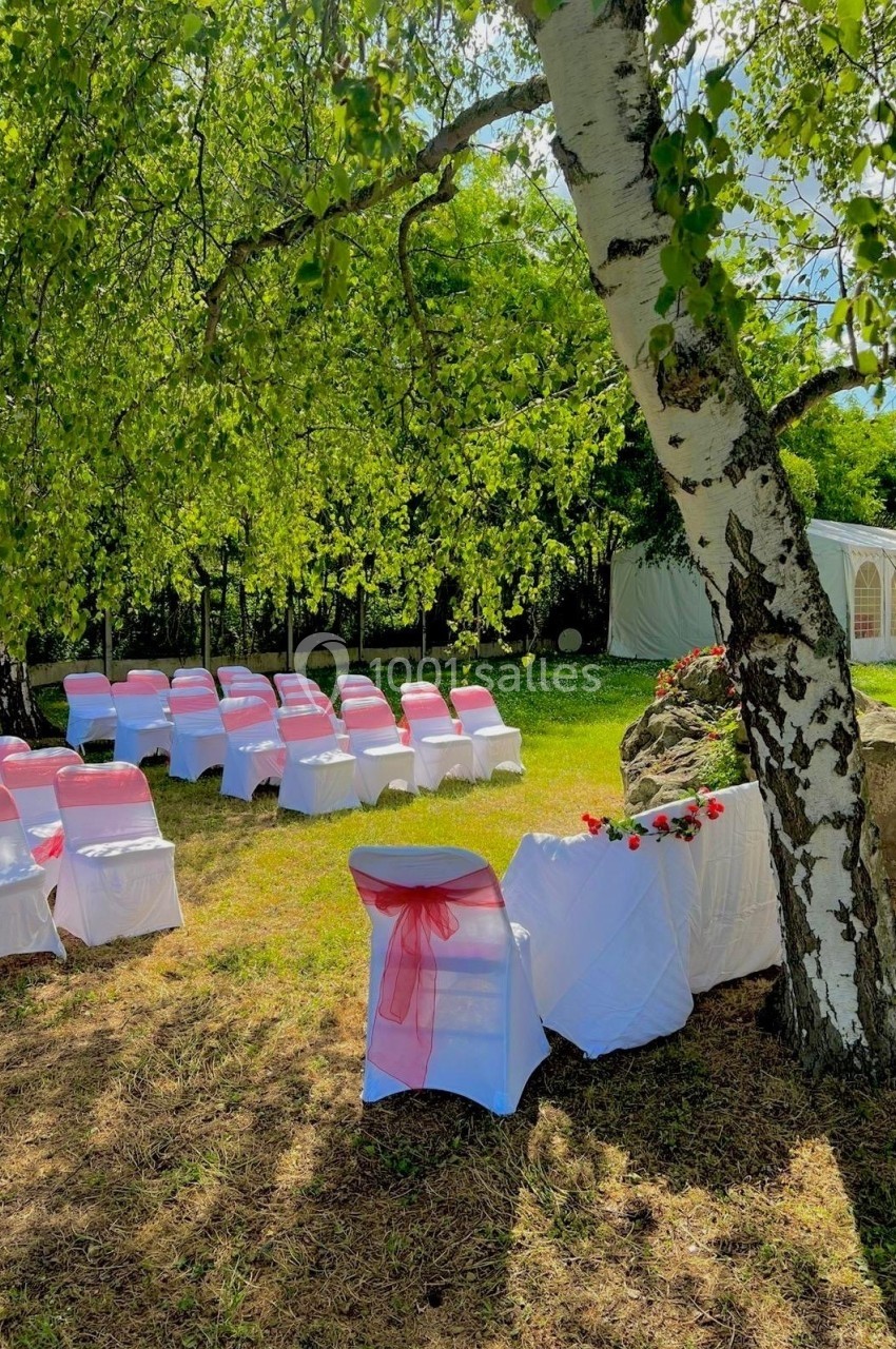 Chaises blanches décorées de nœuds roses disposées en extérieur sous des arbres pour une cérémonie en plein air.