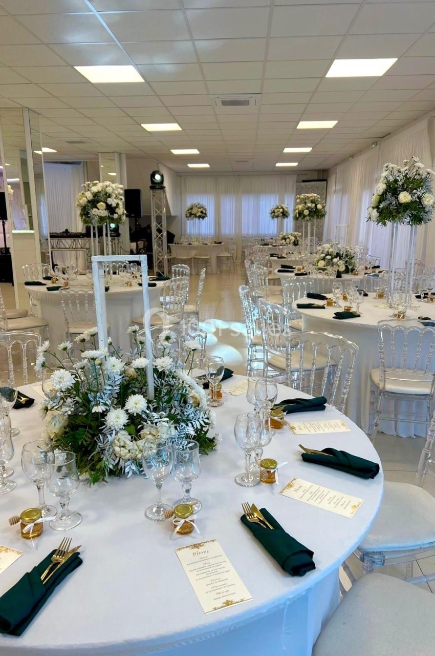 Salle de réception décorée avec des tables rondes, nappes blanches, centres de table fleuris et chaises transparentes.