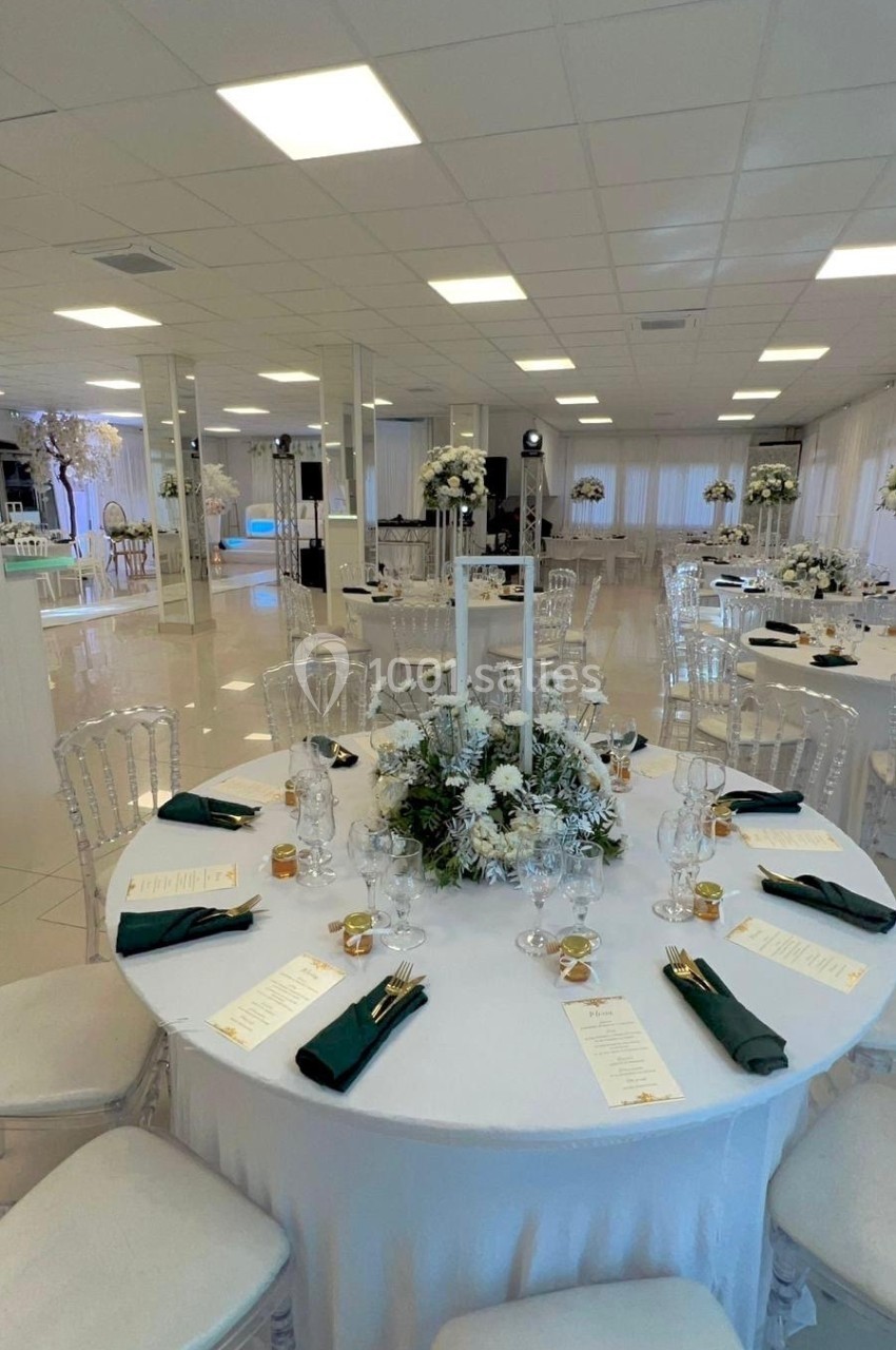Salle de réception lumineuse avec tables rondes décorées de centres de table floraux, nappes blanches et serviettes vertes.