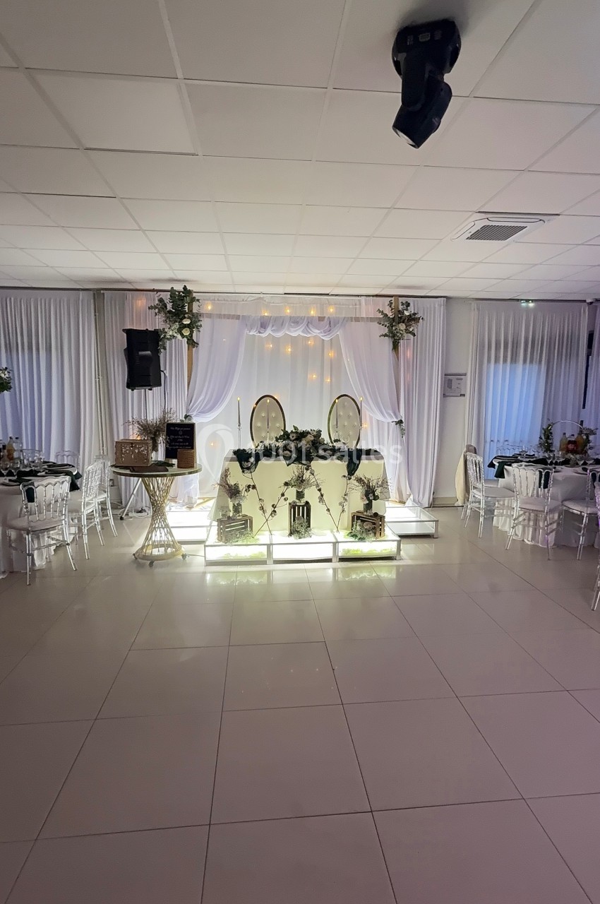 Salle de réception décorée avec une table d'honneur illuminée, des chaises blanches et des rideaux blancs en arrière-plan.