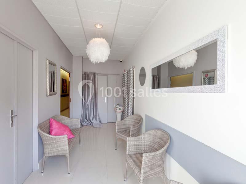 Espace d'attente lumineux avec fauteuils en rotin, coussin rose, miroir mural et décoration suspendue blanche.