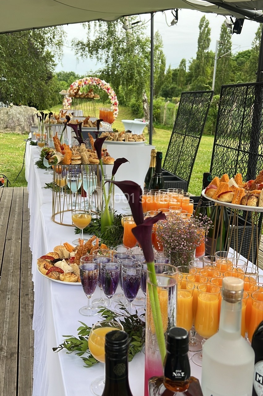 Buffet en plein air avec boissons, viennoiseries, fruits et décorations florales sous une tente dans un jardin verdoyant.