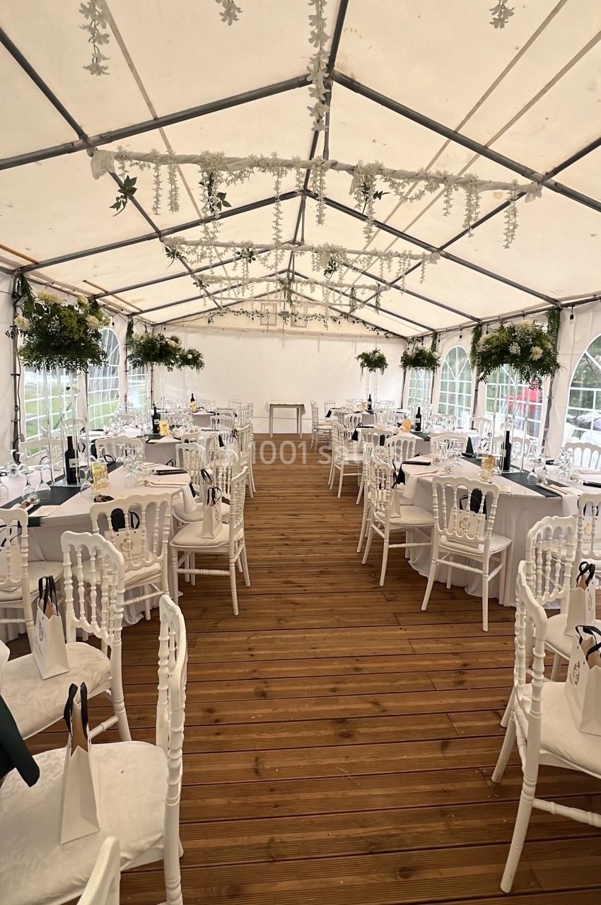 Salle de réception sous une tente blanche, avec tables dressées, chaises blanches et décorations florales suspendues.