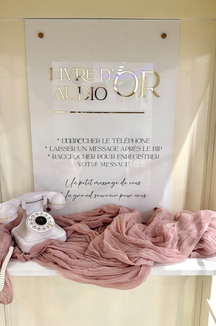 Téléphone rétro blanc posé sur une table avec un tissu rose, devant un panneau indiquant ’Livre d'or audio’.