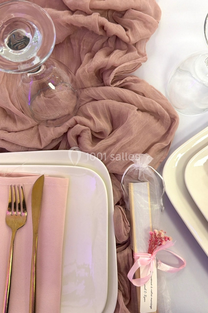 Table dressée avec une assiette blanche, des couverts dorés, une serviette rose, un verre et un cadeau emballé avec un ruban…