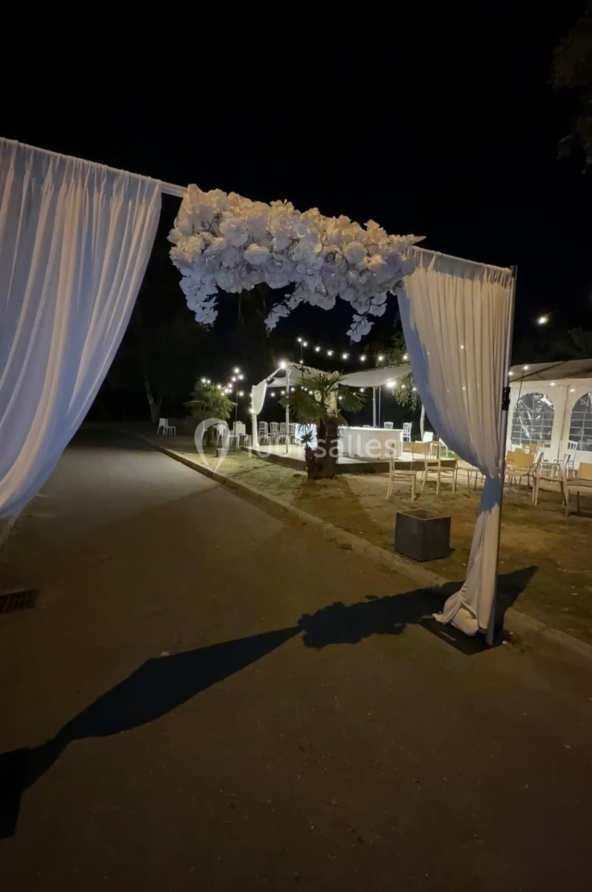 Arche décorée de fleurs blanches et de rideaux, installée en extérieur pour un événement nocturne éclairé.
