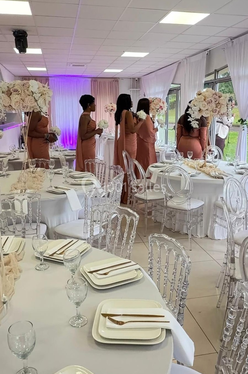 Des femmes en robes roses discutent dans une salle de réception décorée de tables élégantes et de fleurs blanches.