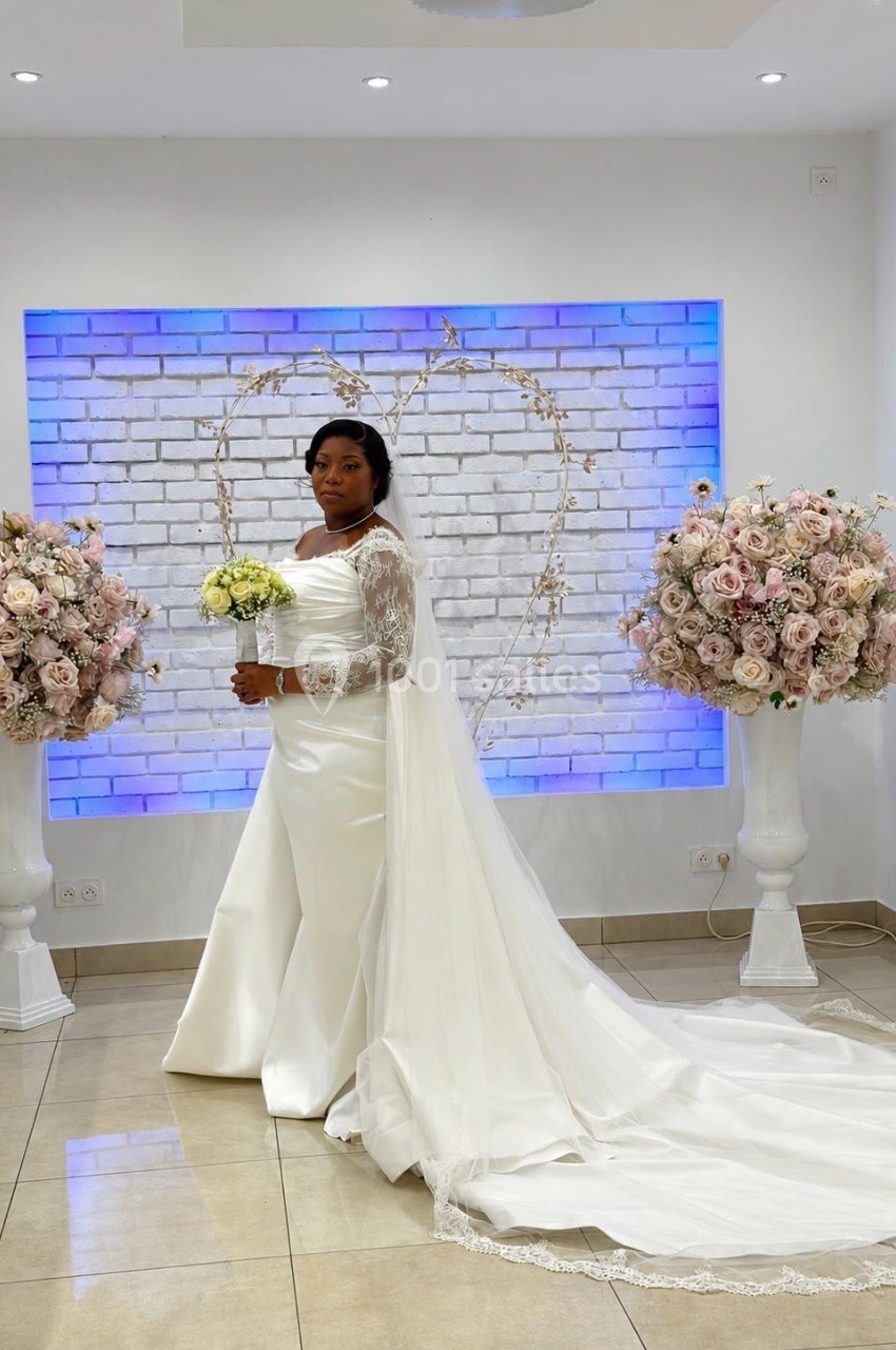 Femme en robe de mariée blanche avec un bouquet, posant devant un mur en briques décoré d'un cœur lumineux.