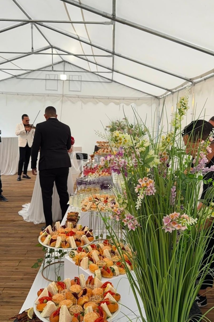 Buffet de desserts variés sur une longue table décorée de fleurs, dans une tente avec des invités en arrière-plan.