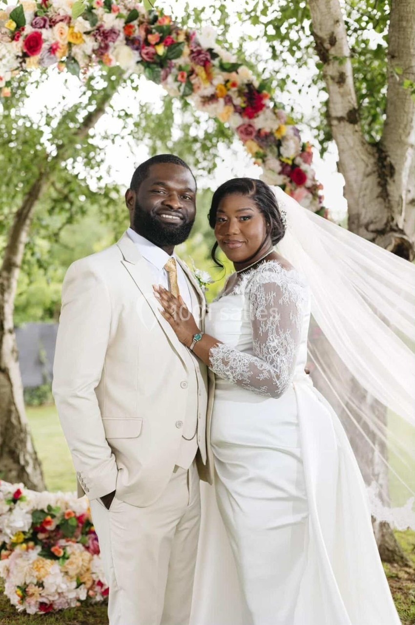 Un couple en tenue de mariage pose devant un arc fleuri sous des arbres dans un cadre extérieur verdoyant.