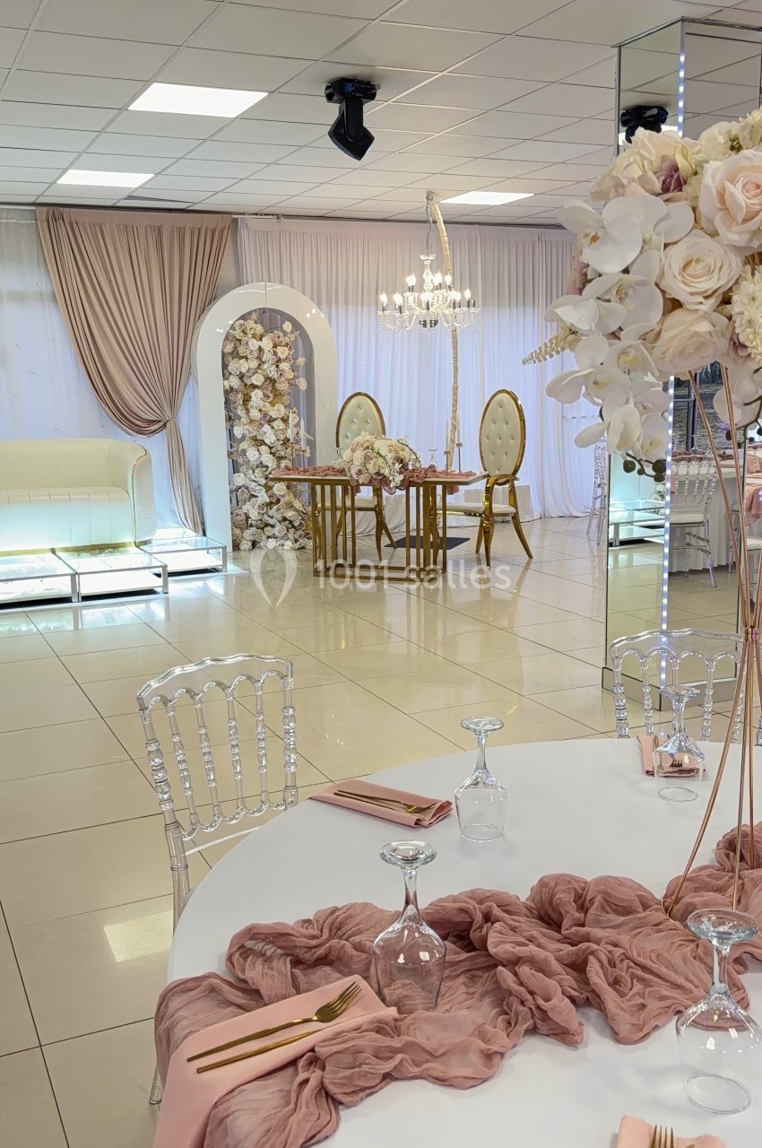 Salle élégante décorée pour un événement avec table dressée, fleurs roses et arche florale en arrière-plan.