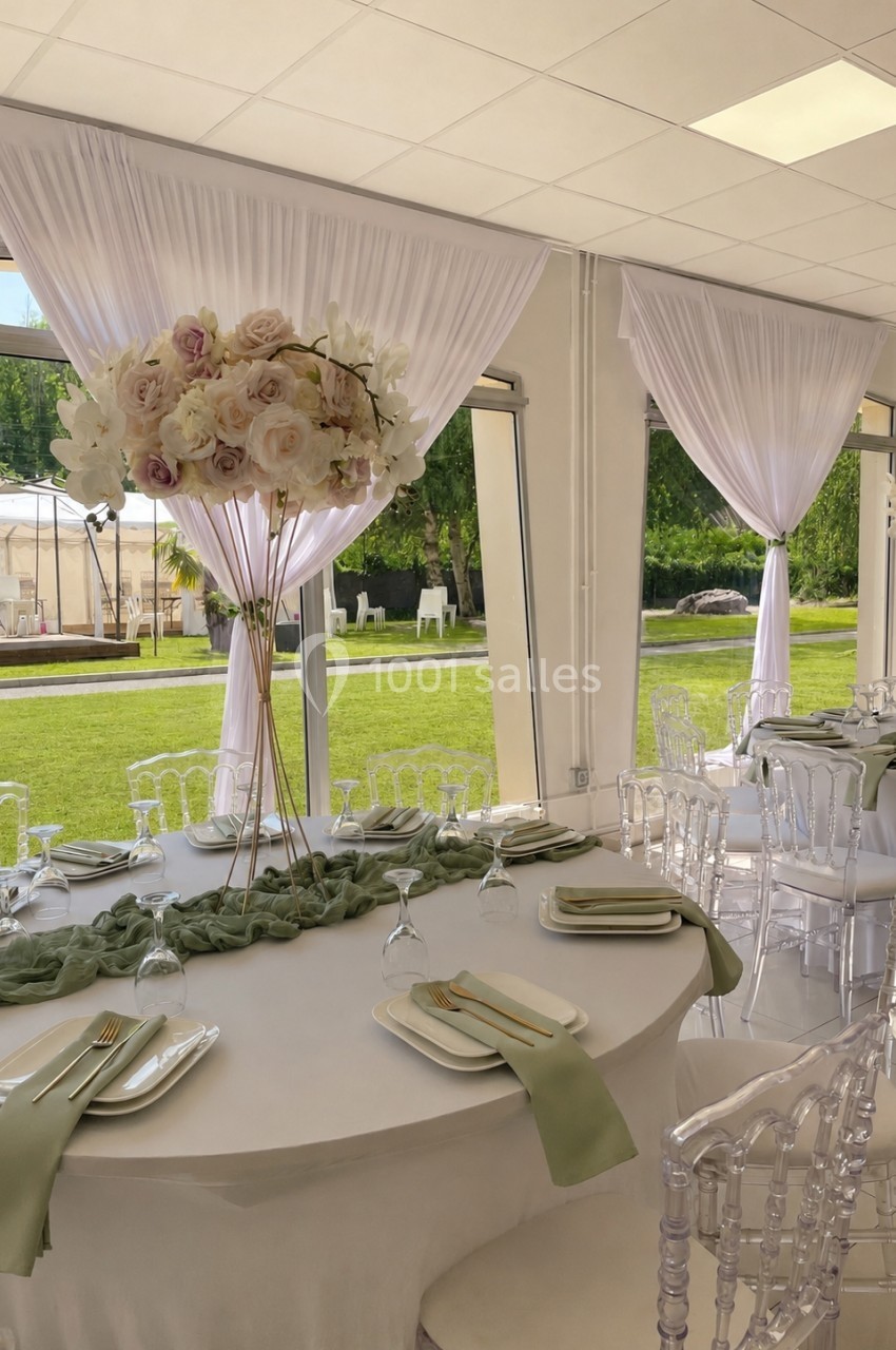 Salle de réception lumineuse avec tables décorées de nappes blanches, serviettes vertes et centres de table fleuris.