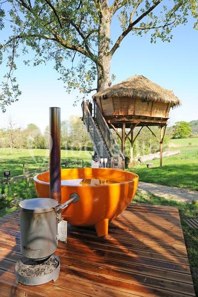 Bain nordique orange sur une terrasse en bois, avec une cabane perchée dans un arbre en arrière-plan.
