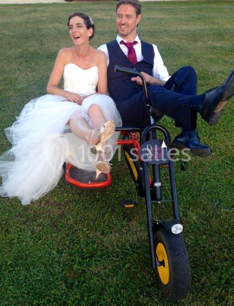 Un couple en tenue de mariage assis sur un tricycle noir dans un jardin.