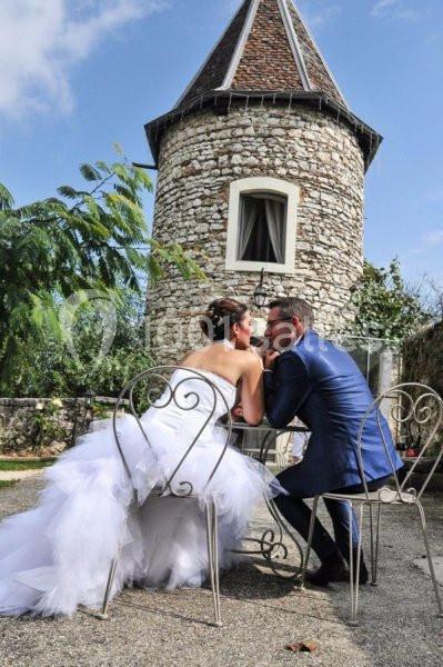 Un couple en tenue de mariage assis à une table en fer forgé devant une petite tour en pierre.