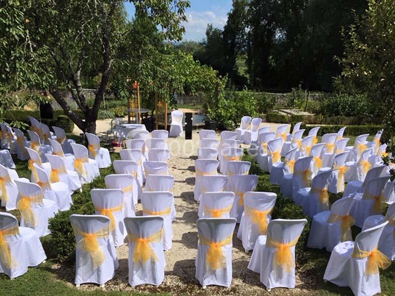 Chaises blanches décorées de nœuds jaunes disposées en rangées dans un jardin pour une cérémonie en plein air.
