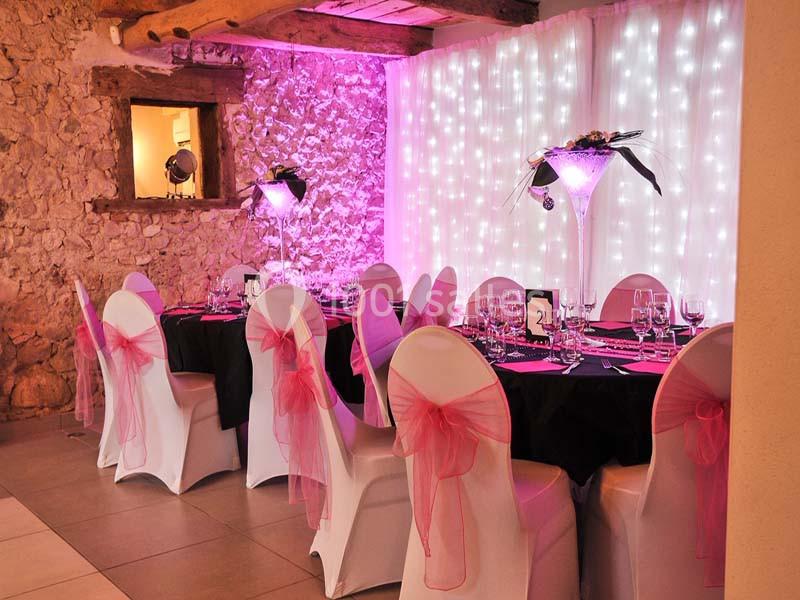 Salle décorée pour un événement avec des tables dressées, des chaises blanches ornées de nœuds roses et un éclairage tamisé.