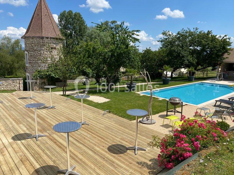Terrasse en bois avec tables hautes, piscine, jardin arboré et tour en pierre sous un ciel bleu.