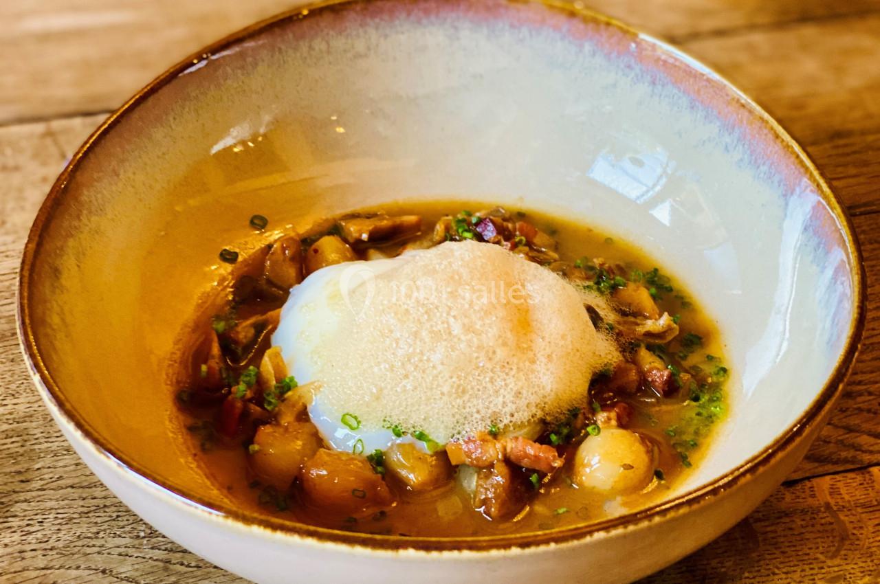 Assiette creuse contenant un plat composé d'un œuf poché, mousse légère, légumes et garniture dans un bouillon.