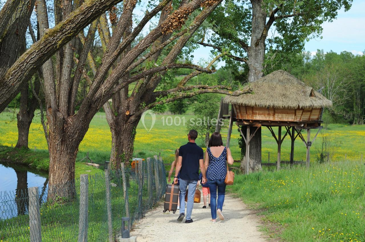 Un couple marche sur un chemin bordé d'arbres, transportant des valises près d'une cabane en bois sur pilotis.