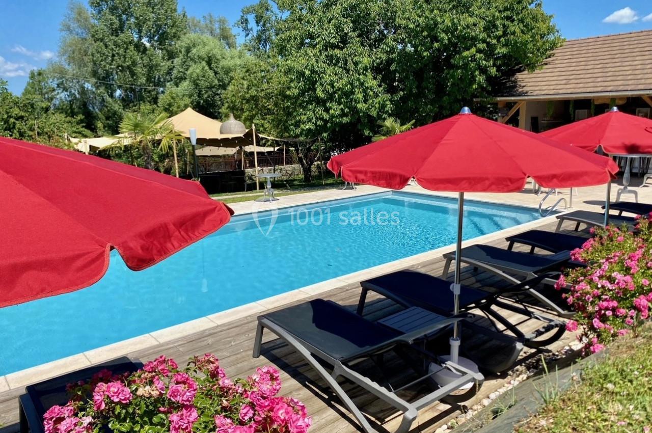 Piscine extérieure entourée de transats noirs, parasols rouges, fleurs roses et végétation verdoyante.