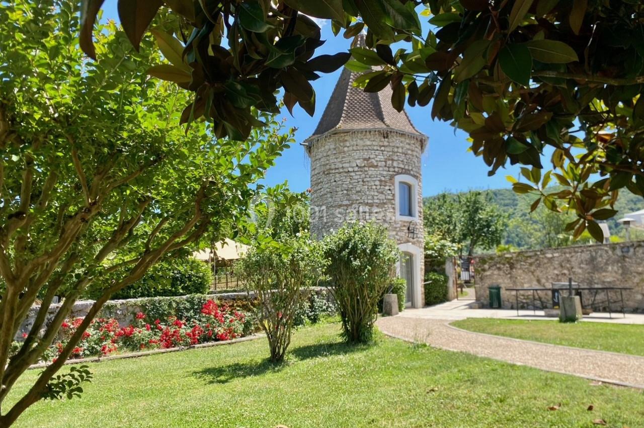 Tour en pierre entourée de verdure, avec des arbres, des fleurs et un chemin dans un jardin ensoleillé.