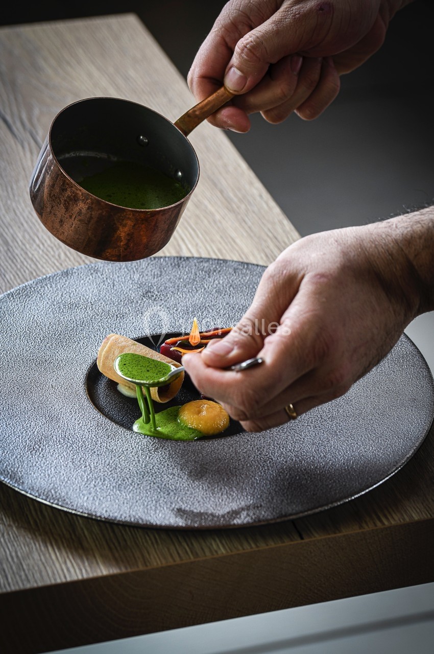 Un chef verse une sauce verte sur un plat raffiné présenté dans une assiette grise texturée.