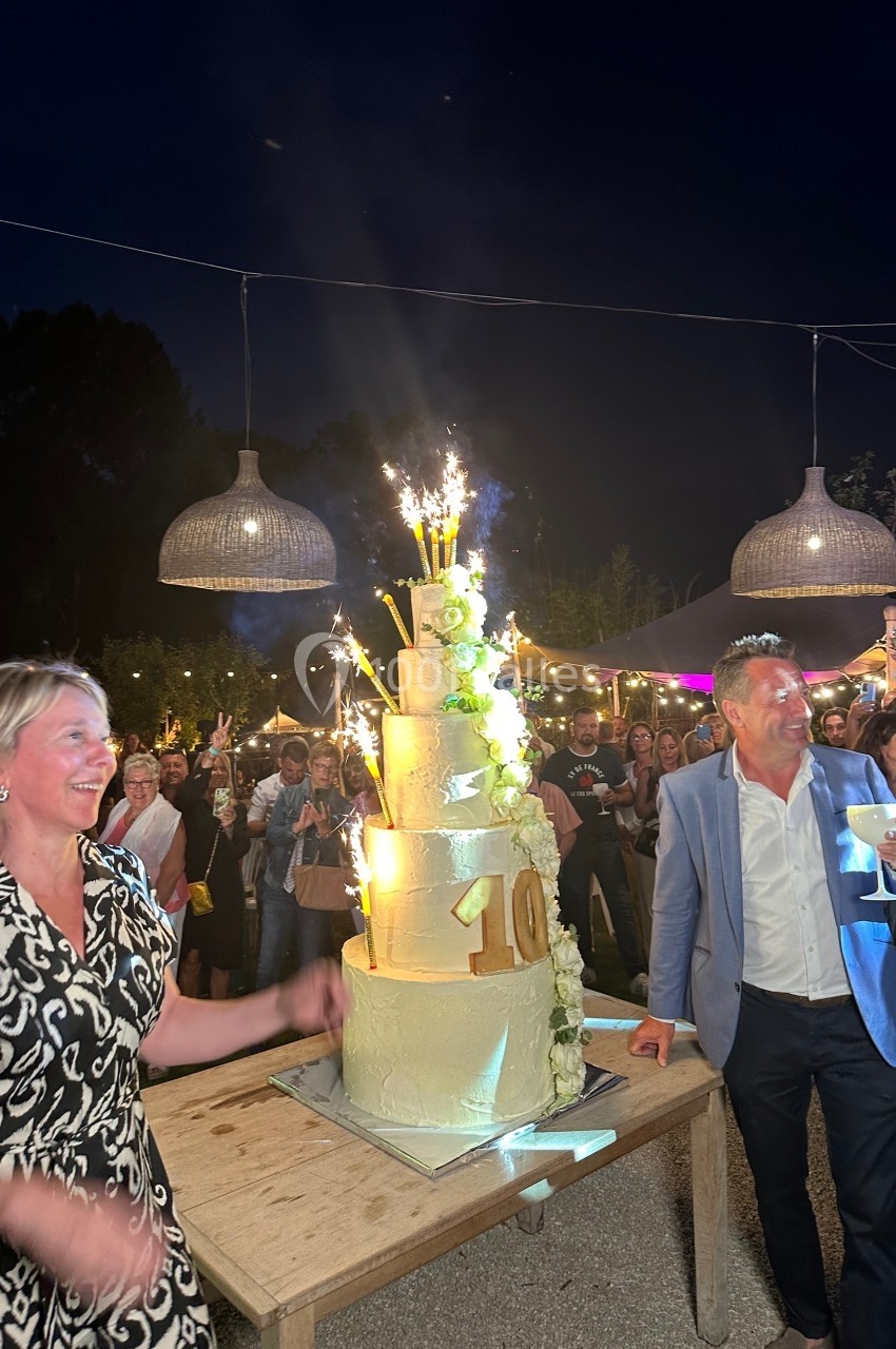 Un gâteau de fête à plusieurs étages avec des bougies étincelantes, entouré de personnes célébrant en extérieur.