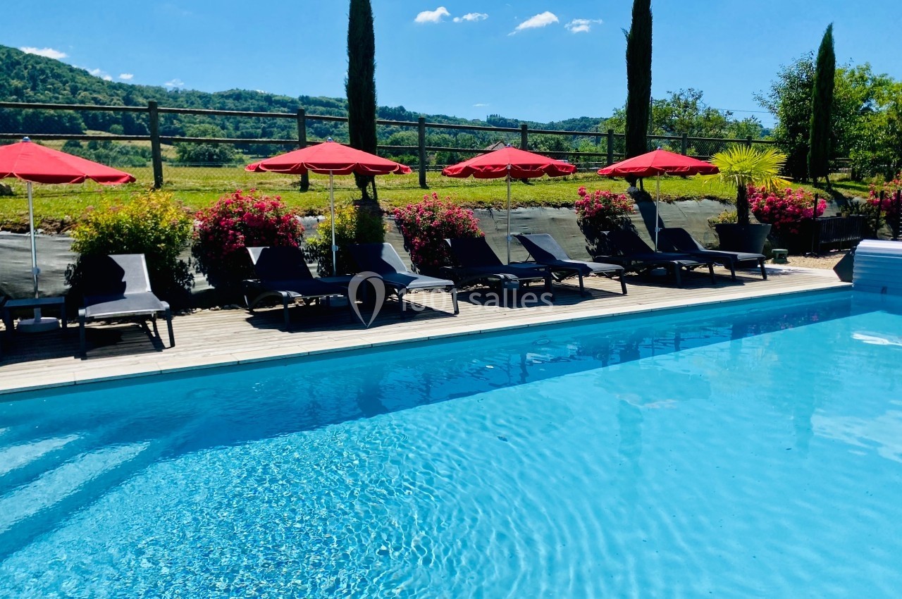 Piscine extérieure entourée de chaises longues, parasols rouges et végétation, avec vue sur des collines verdoyantes.