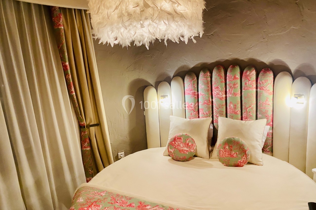 Lit rond avec tête de lit capitonnée à motifs floraux, éclairé par des appliques murales et un lustre à plumes blanches.