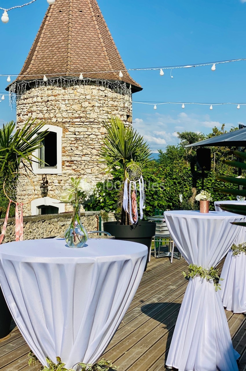 Terrasse ensoleillée avec tables hautes décorées, tour en pierre en arrière-plan et guirlandes lumineuses suspendues.