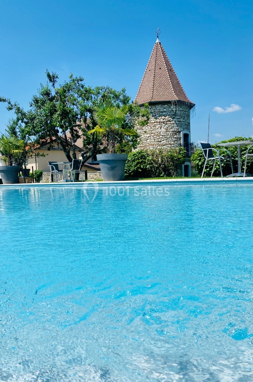Piscine extérieure avec eau turquoise, entourée de chaises, plantes en pots et un bâtiment en pierre avec toit en pointe.
