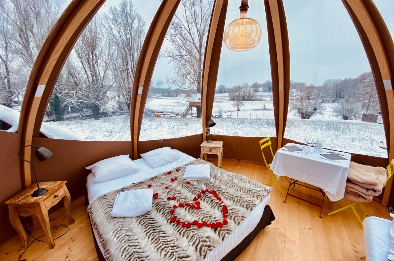 Intérieur d'une bulle en bois avec lit décoré de pétales de rose, vue sur un paysage enneigé à travers de grandes baies…