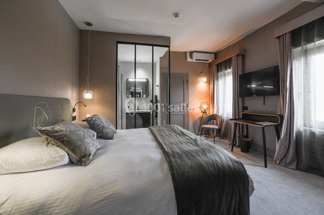 Chambre moderne avec lit double, bureau, télévision murale, miroir et lumière naturelle entrant par une grande fenêtre.