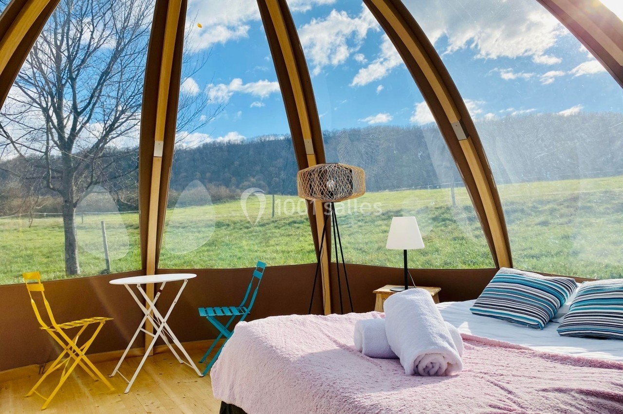 Intérieur d'une bulle avec lit, table et chaises, offrant une vue panoramique sur un paysage champêtre ensoleillé.