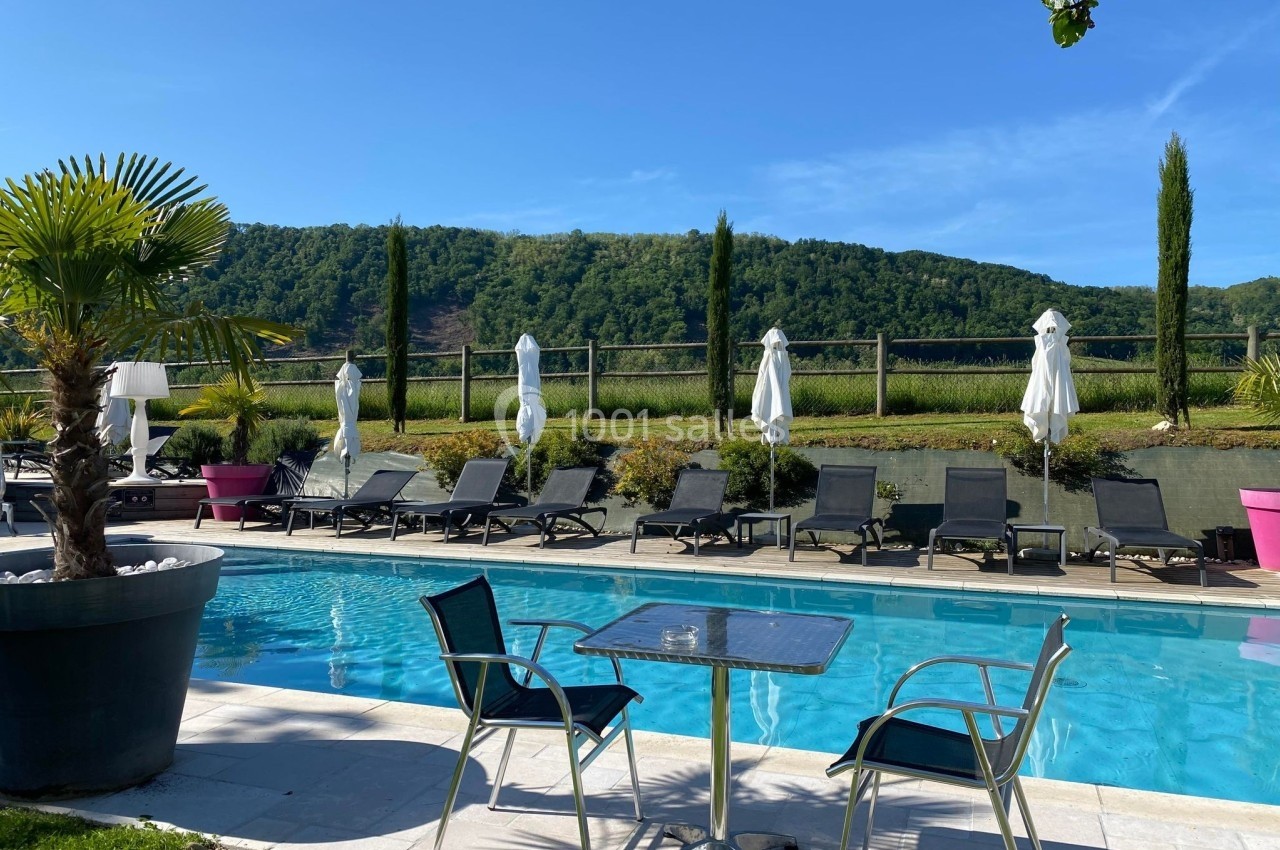 Piscine extérieure avec chaises longues, parasols blancs et table en métal, entourée de verdure et collines.