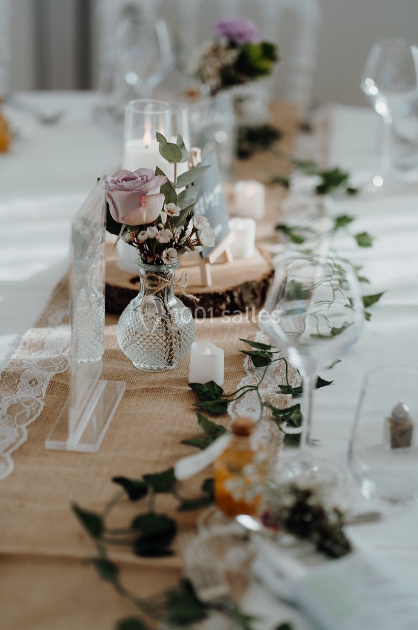 Table décorée avec un chemin de table en toile de jute, des bougies, des fleurs dans des vases en verre et des feuillages.