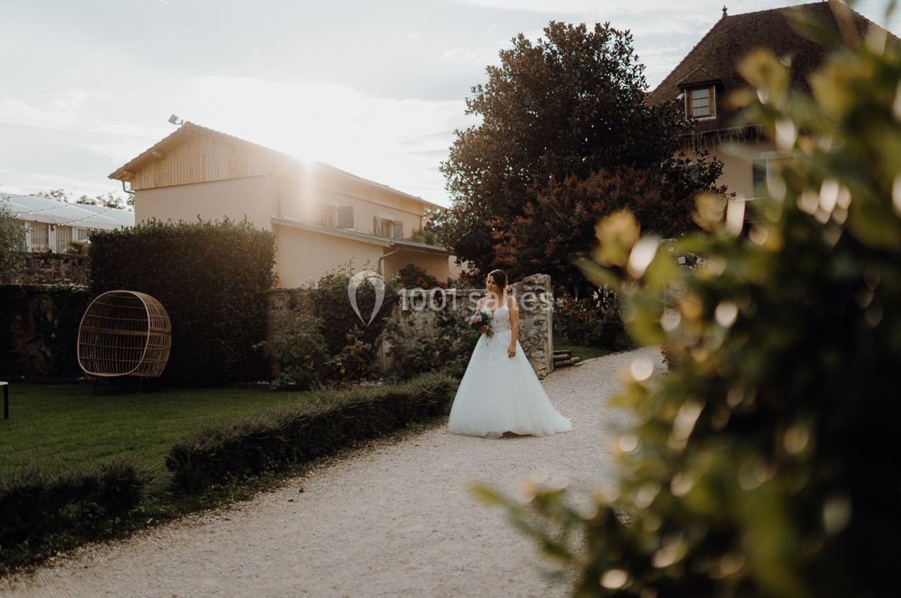 Une mariée en robe blanche marche sur une allée gravillonnée, entourée de verdure et de bâtiments au coucher du soleil.